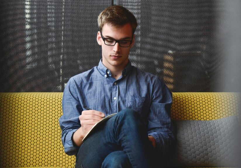 A young man in glasses writes in a notebook while sitting on a stylish couch indoors.