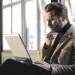 Man looking contemplative while working on a laptop in a well-lit indoor space.