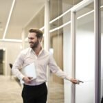 A smiling man holds a laptop while standing in a modern office hallway, exuding confidence.