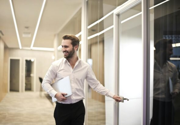 A smiling man holds a laptop while standing in a modern office hallway, exuding confidence.