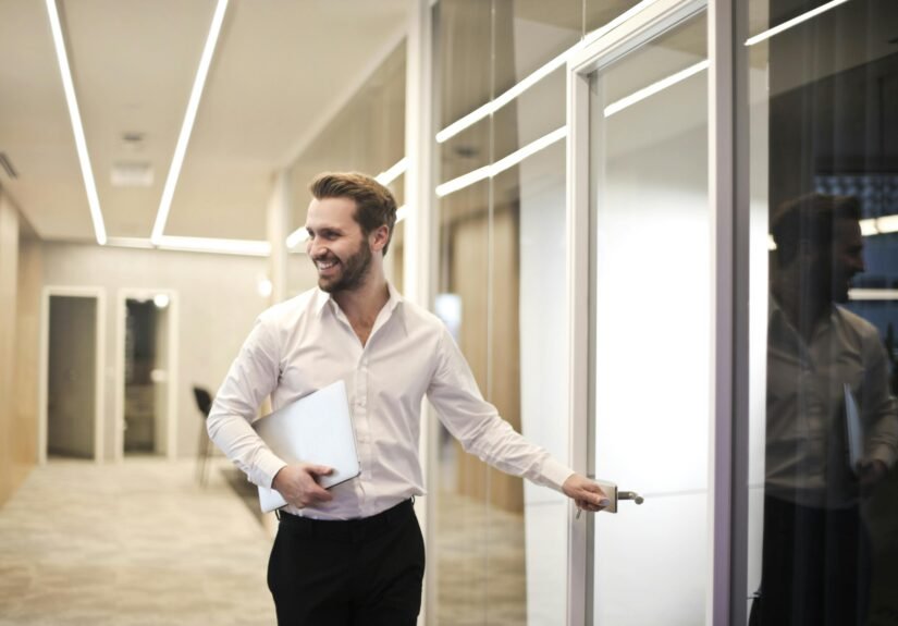 A smiling man holds a laptop while standing in a modern office hallway, exuding confidence.